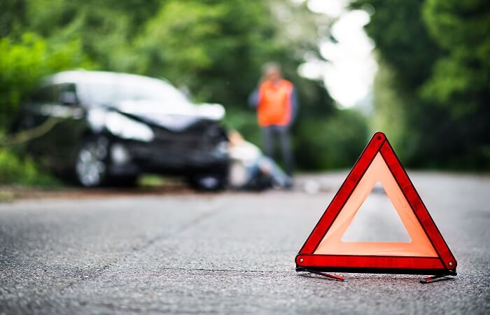 Closeup of a hazard triangle in the middle of the road with an out-of-focus crashed car in the background.