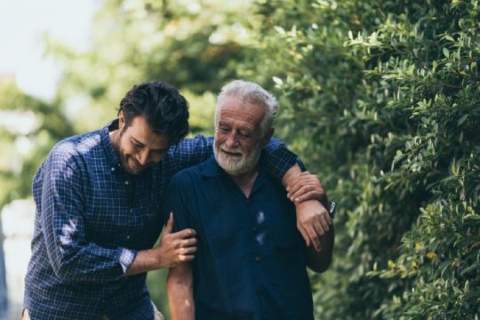 A younger man walks through a treelined area with his arm around the neck and shoulder of an older man.