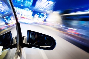 A stylized photo of a side mirror in clear detail in the foreground with the background a blur of time-lapse city lights, indicating the car is speeding